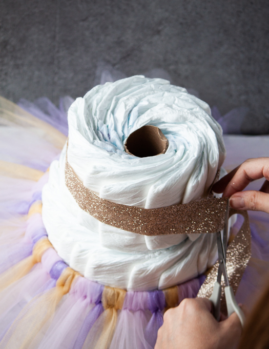 gold ribbon being placed around the top tier of white diaper cake with purple, pink and gold tutu on the bottom tier. 