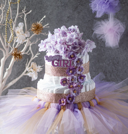 Girls 2 layers diaper cake with pink and purple flowers, gold ribbons, baby girl 3D stickers and pink, gold and purple tutu with pink and purple pom pom, white tree with flowers and gold pom pom and a grey background