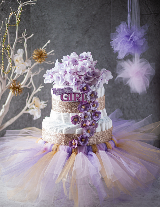 2 tier girls diaper cake decorated with floral, paper flowers, ribbons and a tutu, with pink and purple pom pom, white tree decorated with white flowers and golden pom pom. 