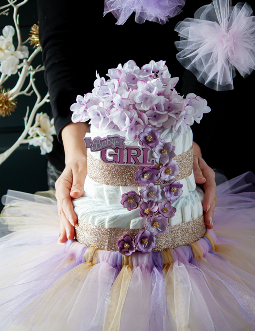 fully decorated diaper cake with pink and purple flowers, gold ribbon and a tutu hold in between 2 hands 