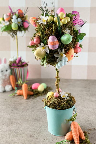 two fully decorated spring topiary with colourful eggs, folders and greenery, carrots and white bunny leaning on the topiary